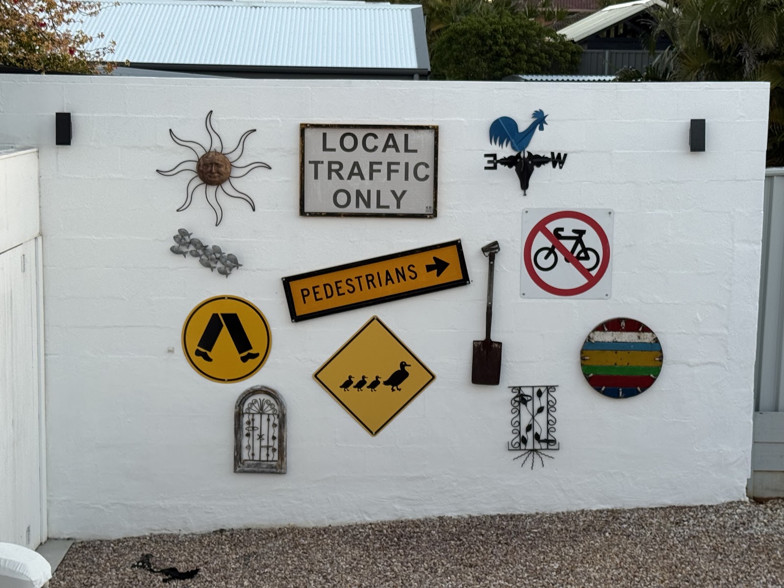 A white rendered driveway wall hung with road signs, a rooster weathervane and metal art